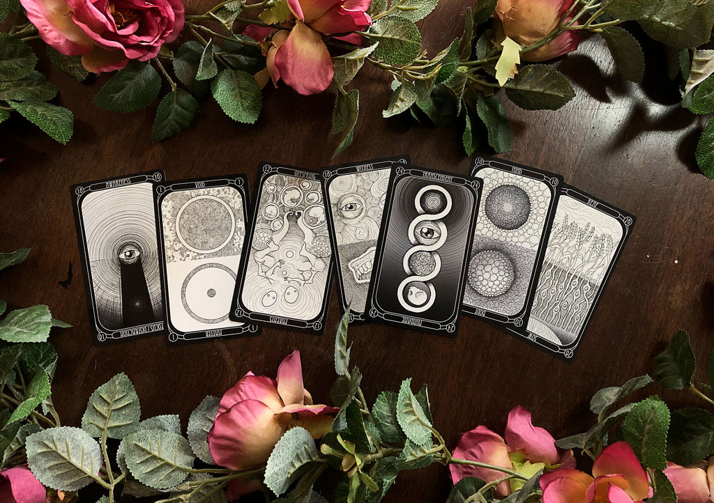 Seven illustrated cards from The Eigengrau Oracle Deck Limited Drop by Rage Create, with intricate black-and-white designs, are displayed on wood amid pink roses and green leaves. This hand-drawn oracle deck features abstract, mystical imagery.