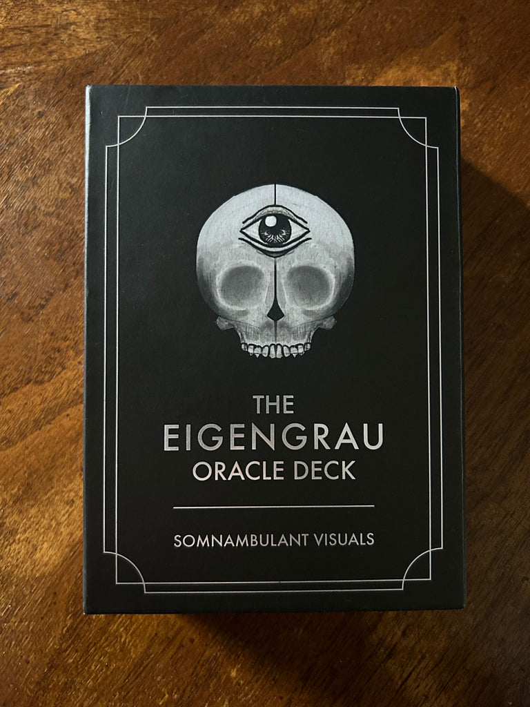 A black box with a skull featuring a third eye is displayed on wood. Below, it reads: “The Eigengrau Oracle Deck Limited Drop” by Rage Create. This hand-drawn oracle deck is from the Somnambulant Visuals series.