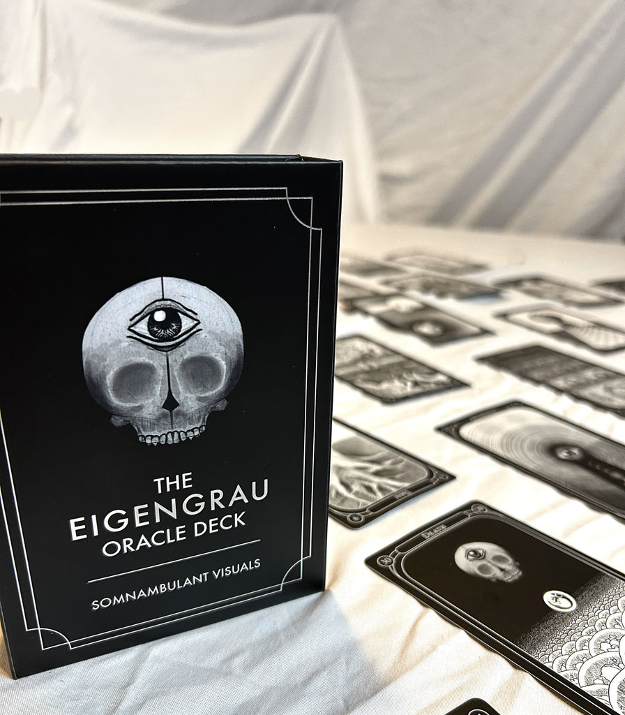 A black box labeled The Eigengrau Oracle Deck Limited Drop by Rage Create, featuring a skull with an eye on its forehead, sits on a table with various hand-drawn, double-sided oracle cards spread out on a white cloth.