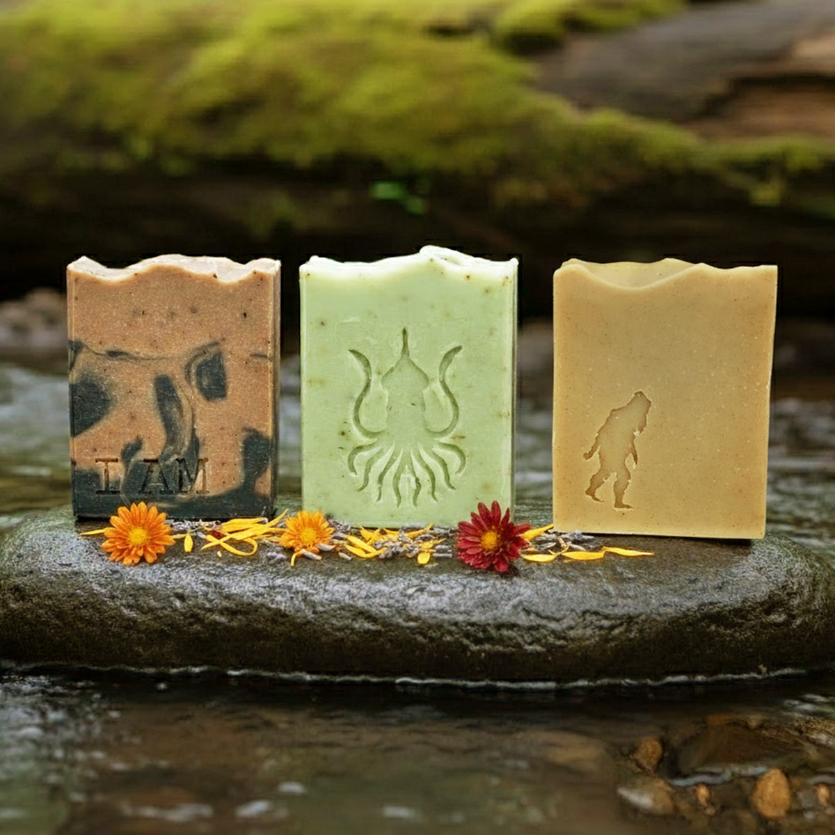 The Rage Create Bar Soap 3-Pack *Limited Drop* by Rage Create features small-batch soaps with bear, squid, and Bigfoot wildlife designs on a stone by a stream, accented with yellow and orange flowers against moss and log in the blurred background.