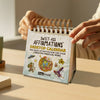 Sweet-Ass Affirmations Desktop Calendar held by a person on a wooden surface