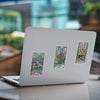 Three colorful tarot-style sticker cards featuring a sea turtle, bison, and red panda with intricate rainbow patterns and ornate borders, arranged on a silver laptop lid in a garden setting.