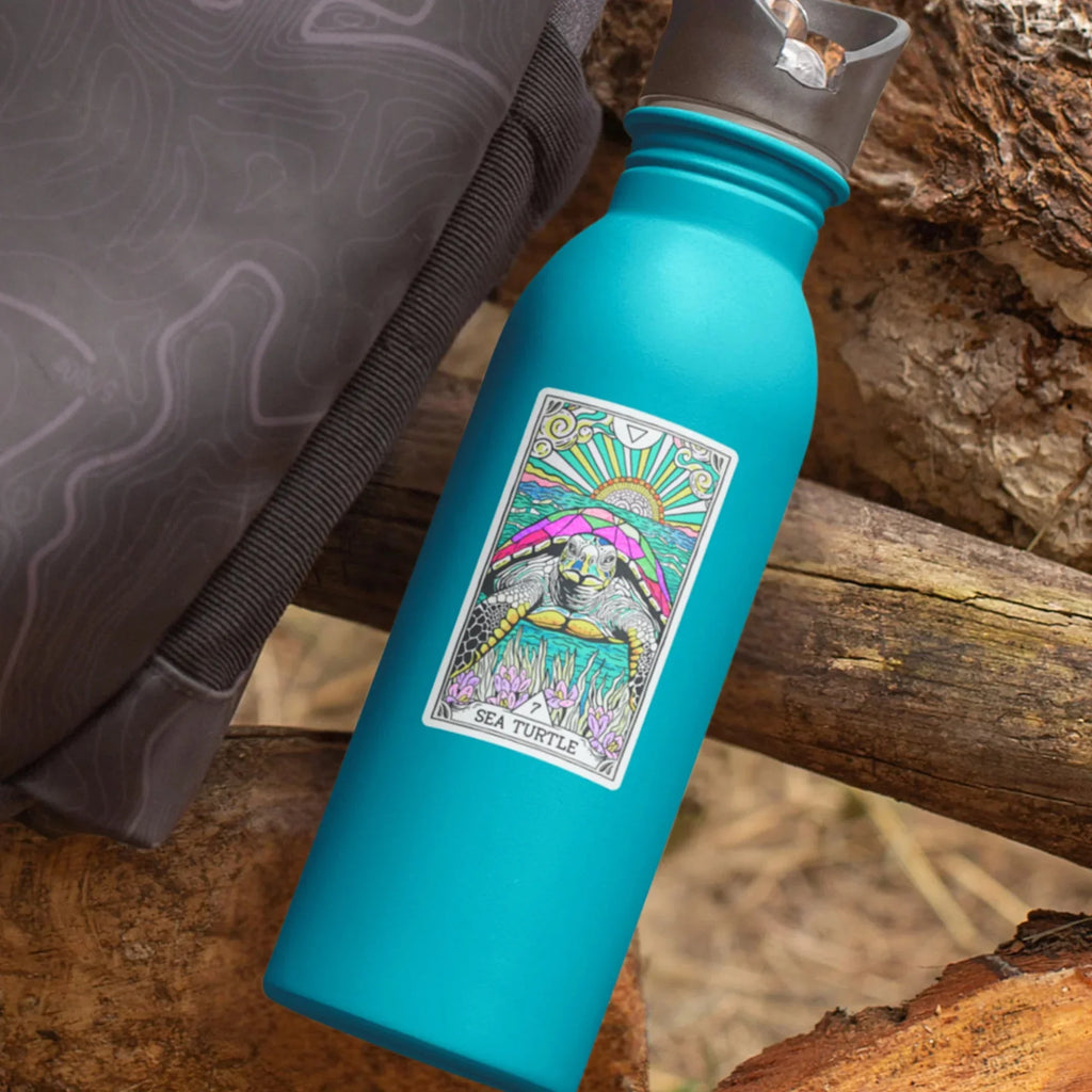 Bright turquoise water bottle with a colorful Sea Turtle tarot-style sticker label, placed on weathered driftwood and sand with a dark patterned fabric nearby.