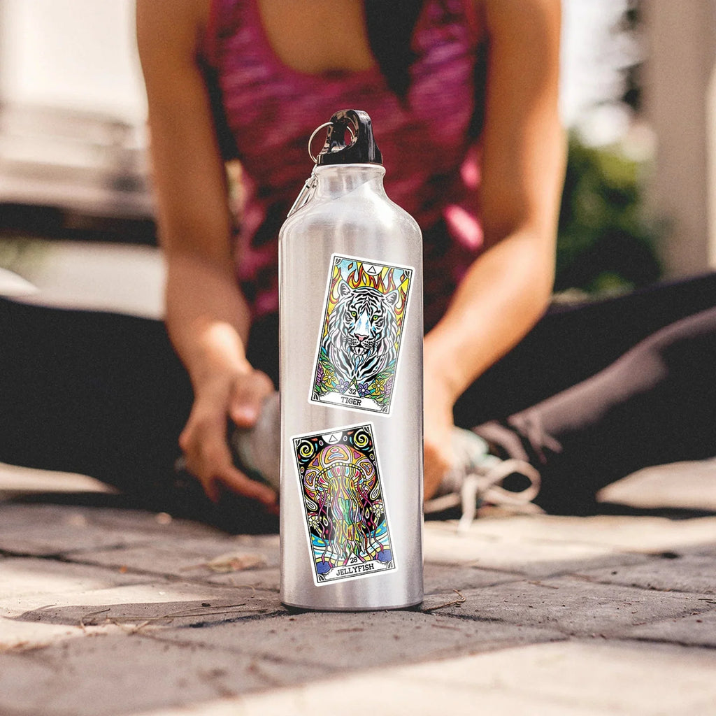 White aluminum water bottle featuring vibrant oracle card stickers with tiger and jellyfish designs, held by a person in a magenta tank top outdoors on pavement.