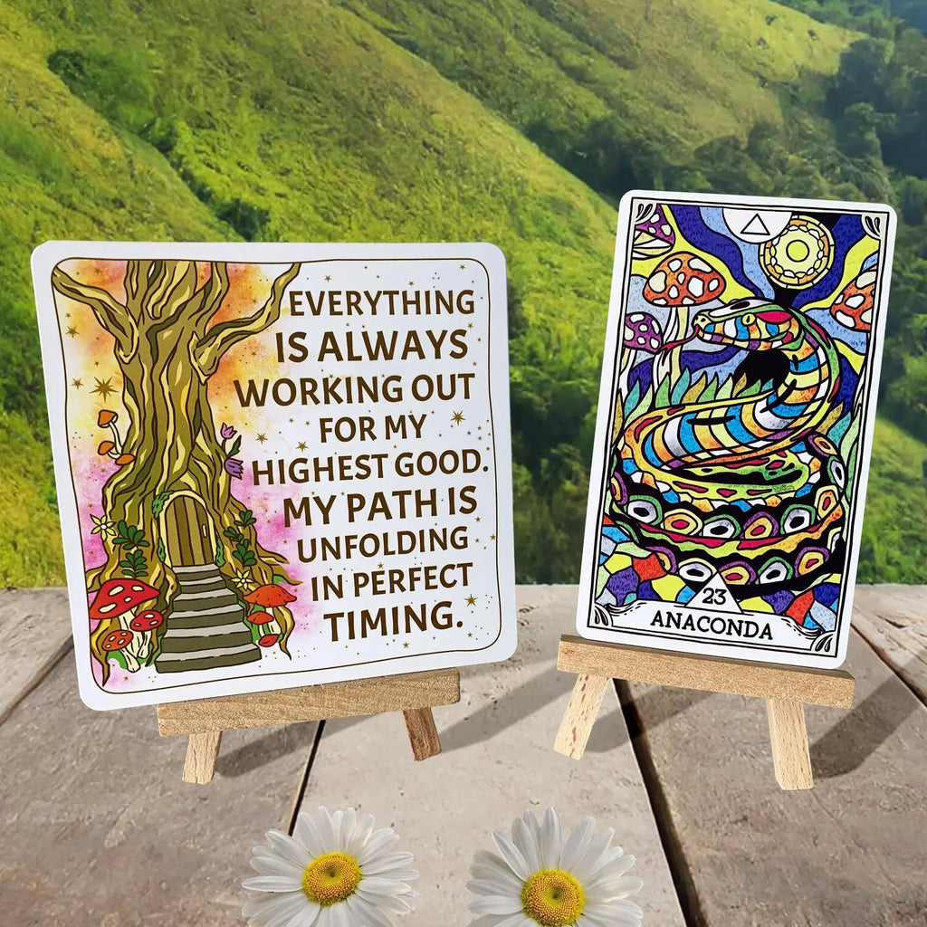 Two wooden easel card stands displaying an affirmation card with a whimsical tree door and an Anaconda oracle card with vibrant psychedelic patterns, set on weathered wood with white daisies and lush
