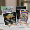 Three wooden easel stands displaying colorful affirmation and oracle cards, including a black card with a stegosaurus and motivational text, alongside mystical tarot imagery with rainbow and mushroom