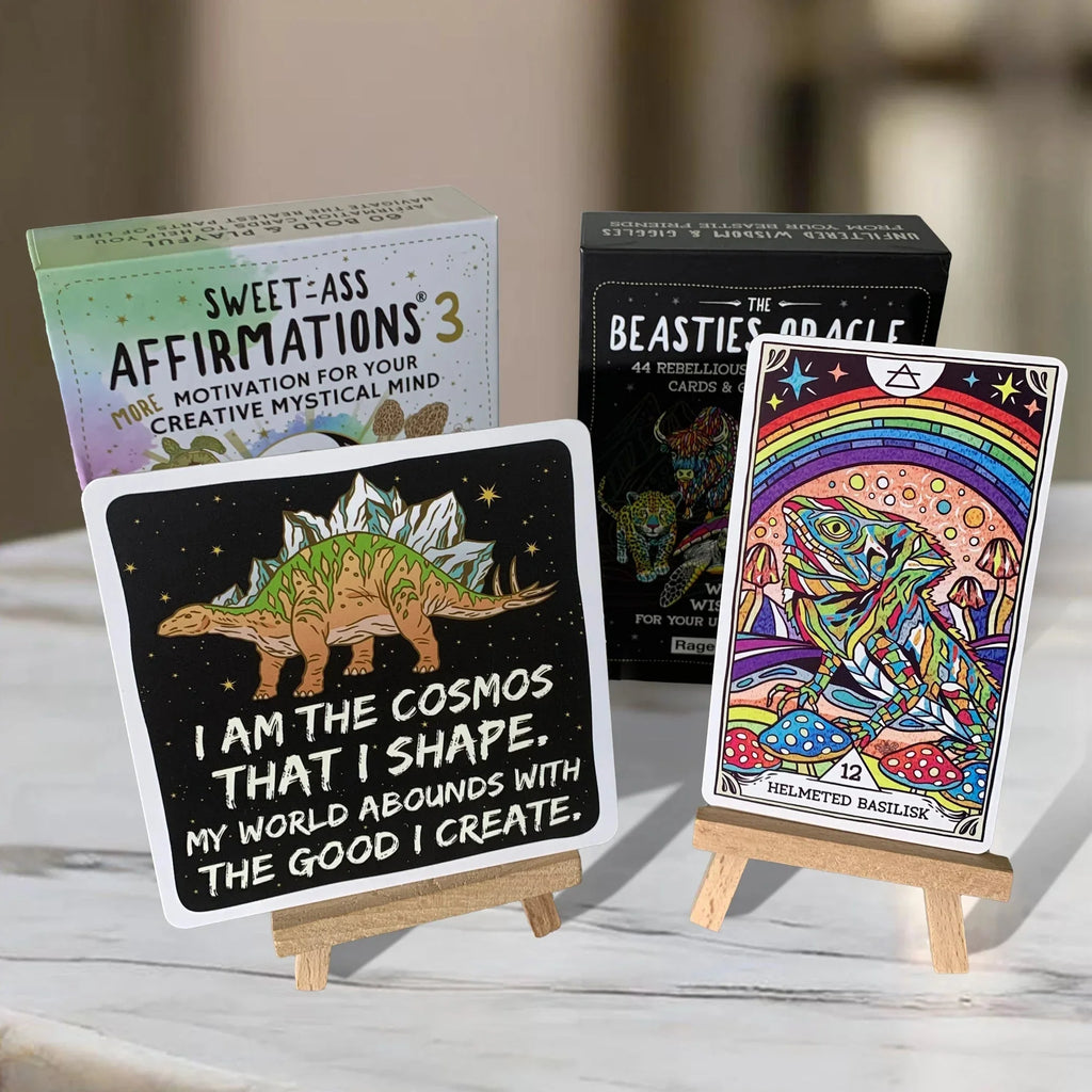 Three wooden easel stands displaying colorful affirmation and oracle cards, including a black card with a stegosaurus and motivational text, alongside mystical tarot imagery with rainbow and mushroom