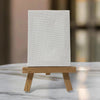 A blank white textured canvas sits on a small wooden tripod easel against a soft, blurred beige and green background, ready for custom art or affirmations.