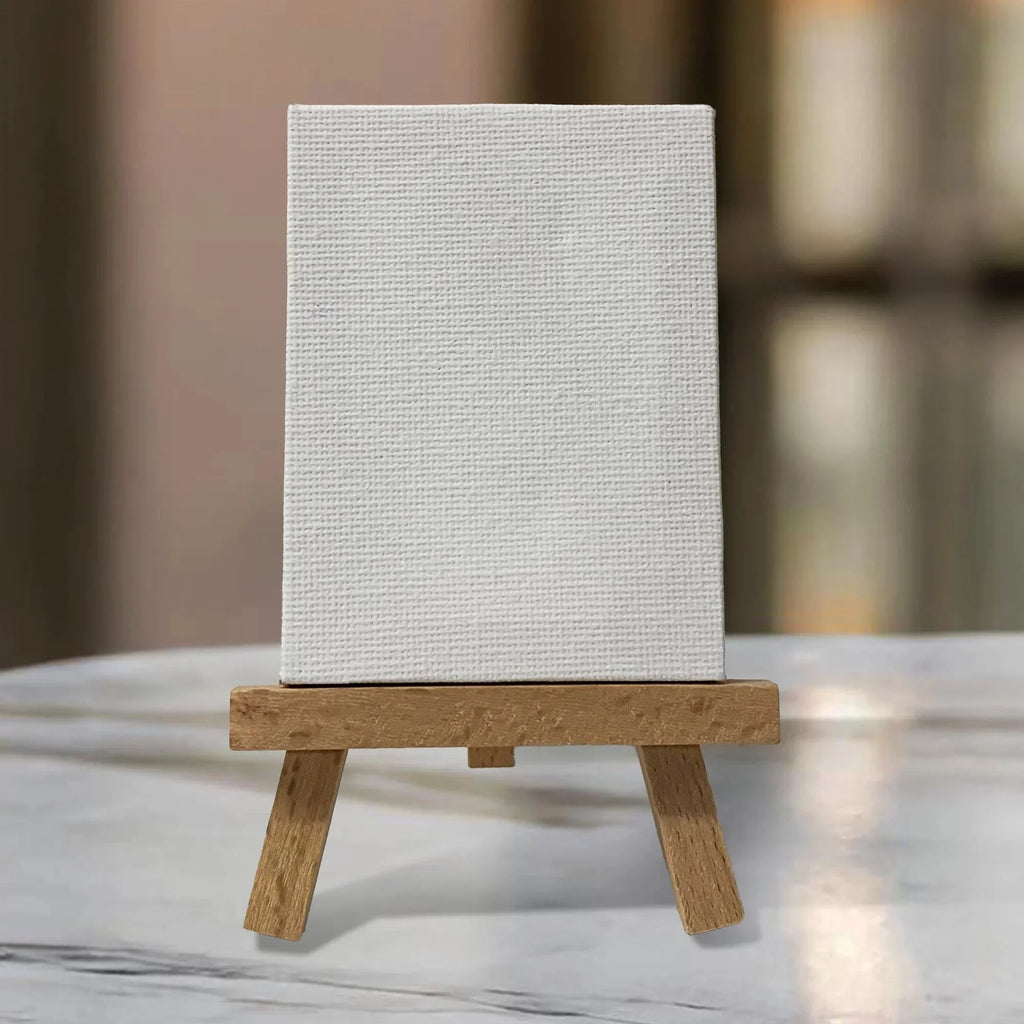 A blank white textured canvas sits on a small wooden tripod easel against a soft, blurred beige and green background, ready for custom art or affirmations.