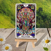 Colorful jellyfish tarot card numbered 28 displayed on a natural wood easel with white daisies on weathered wood deck and green foliage backdrop.