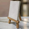 White textured canvas card sits on a wooden easel stand with natural wood legs, photographed from a front angle on a light surface with warm sunlight casting shadows.