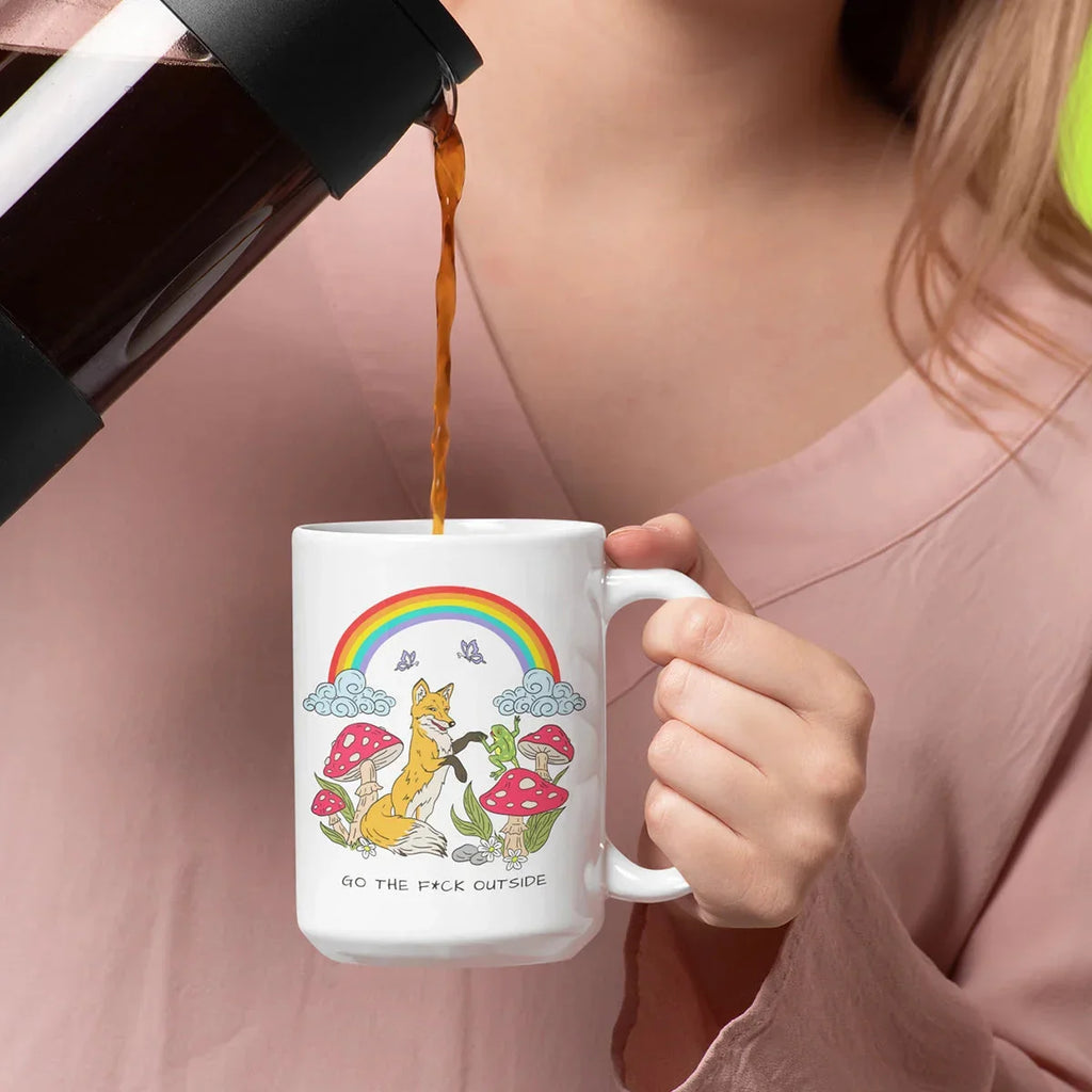 Person in pink clothing pouring amber coffee into a white mug featuring a whimsical rainbow, fox, mushrooms, and clouds illustration with bold text.