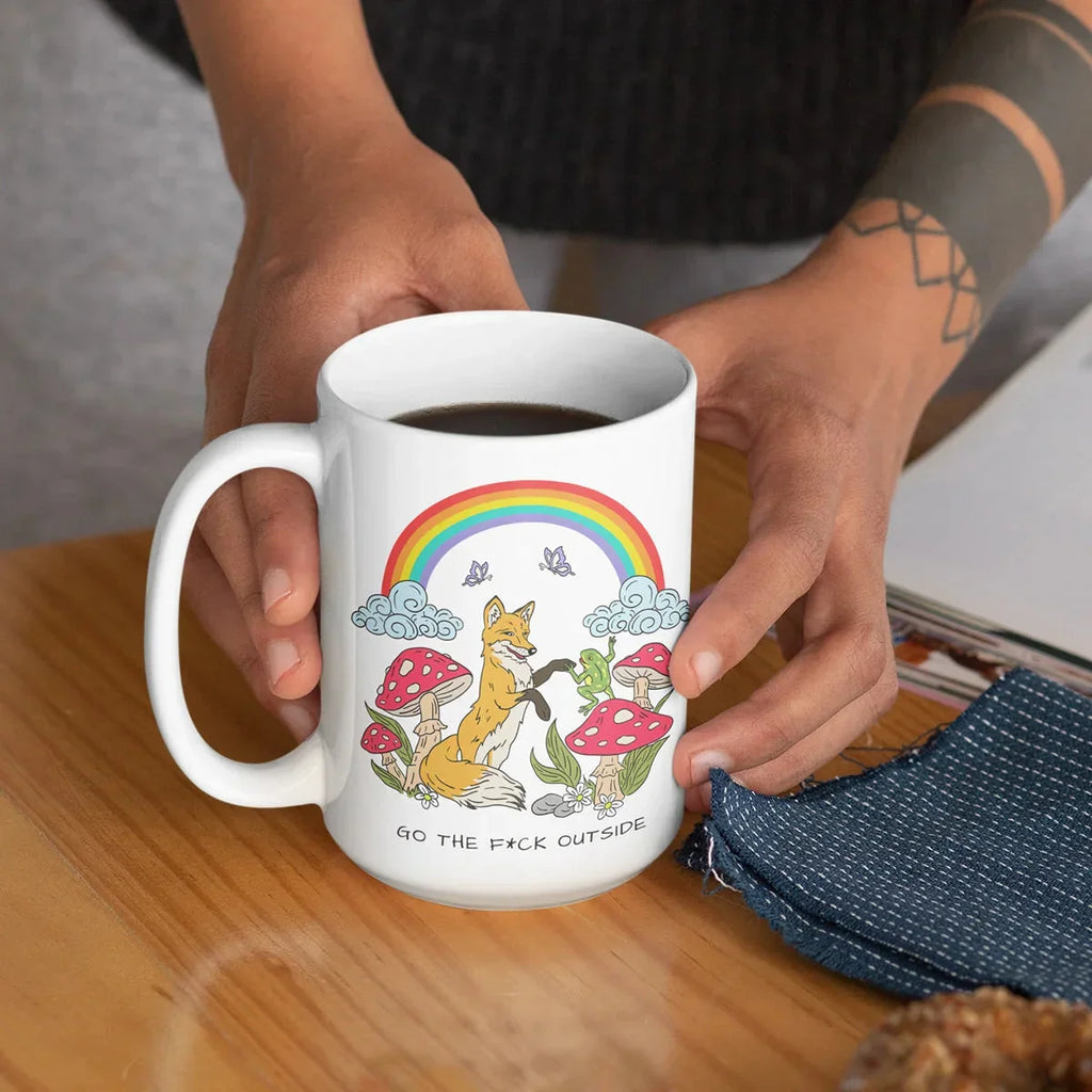 Person with a geometric tattoo holding a white ceramic mug featuring a fox, rainbow, mushrooms, and clouds with the text 'Go the F*ck Outside' while seated at a wooden table.