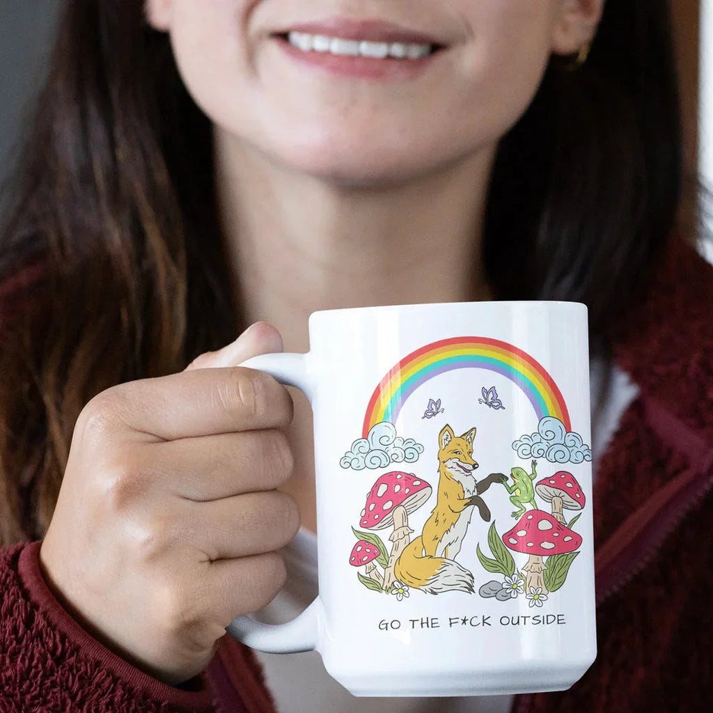 Person in maroon sweater holds a white ceramic mug featuring an illustration of a fox surrounded by red mushrooms, colorful rainbow, clouds, and butterflies with text reading 'GO THE F*CK OUTSIDE'.