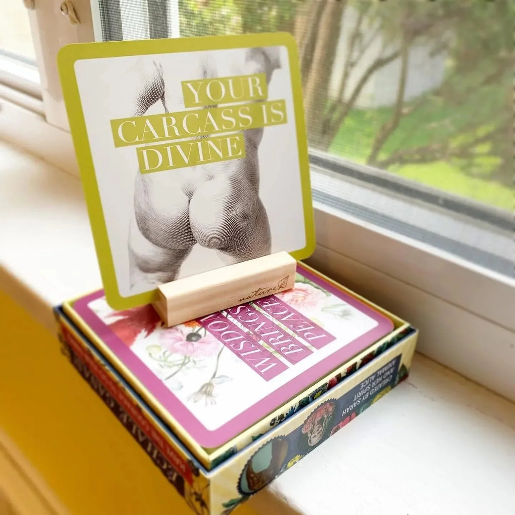 An opened deck box with a lime-green lid featuring an illustration of a sculptural torso and the text 'Your Carcass is Divine,' displayed on a windowsill with pink card pages visible inside and green