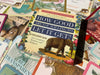 Sarah Bamford Seidalmann's How Good Are You Willing to Let It Get? card deck box displayed flat among other books and manuscripts, featuring cream and navy cover design with floral borders and an illu