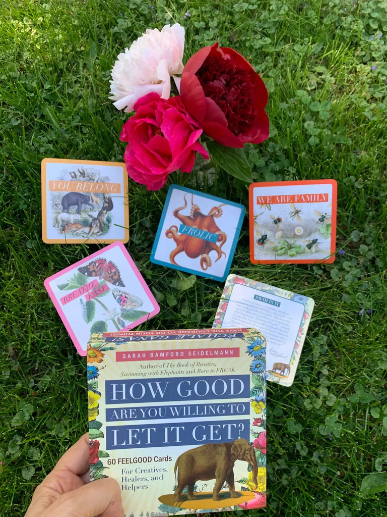 Sarah Bamford Sidelemann's affirmation card deck displayed on green grass with pink, red, and white peonies above, showing colorful card box covers and instruction cards in a flat-lay arrangement.