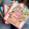 Stack of affirmation cards with pink and teal borders fanned across blue fabric, featuring a profile illustration and colorful collage artwork on the card faces.