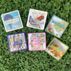 Six colorful affirmation cards from the deck arranged on bright green clover ground, featuring diverse imagery including hot air balloon, butterflies, feathers, and nature scenes in blue, purple, pink