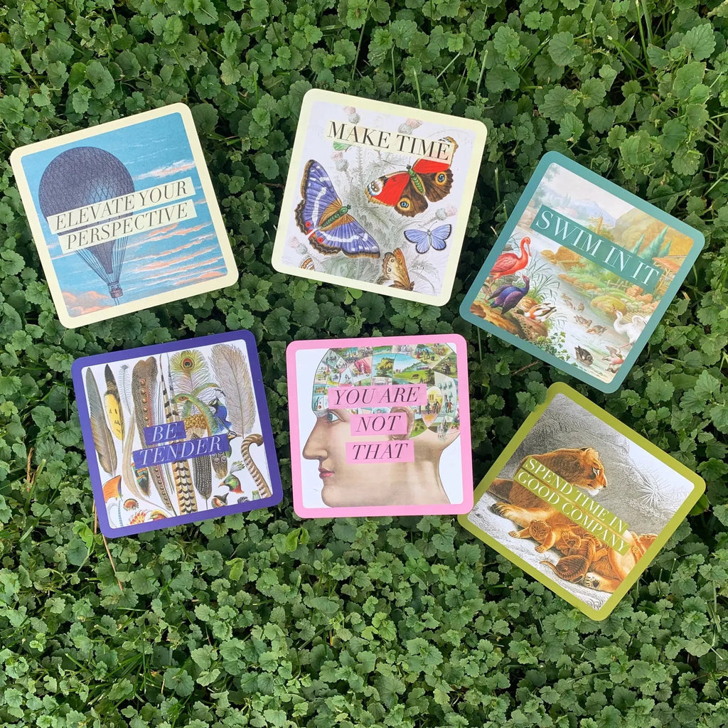 Six colorful affirmation cards from the deck arranged on bright green clover ground, featuring diverse imagery including hot air balloon, butterflies, feathers, and nature scenes in blue, purple, pink
