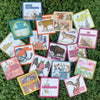 Colorful affirmation cards from the deck arranged in a circular spread on vibrant moss-green grass, showing botanical illustrations, animal imagery, and inspirational text in various colored borders i