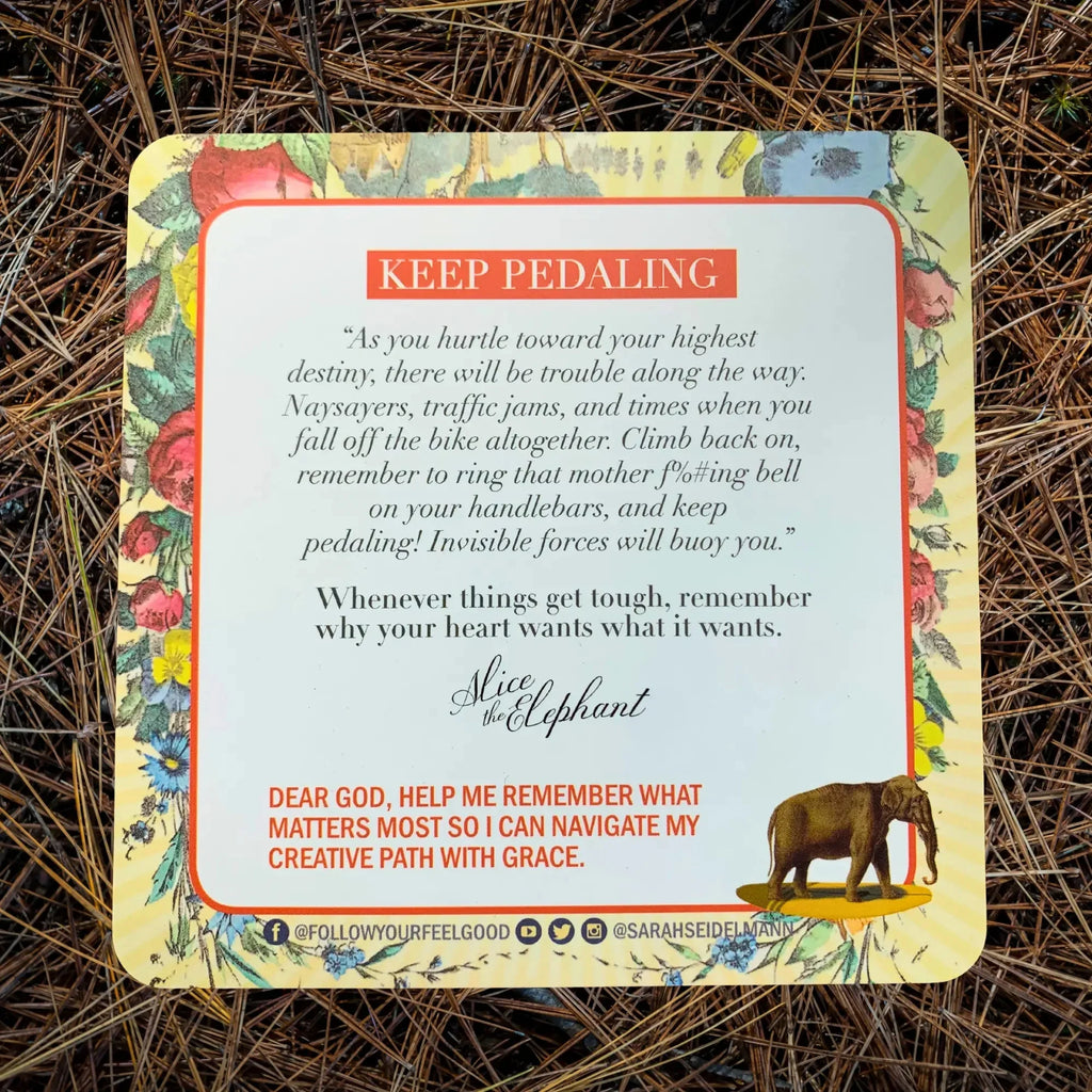 An inspirational card with floral borders and an elephant illustration displays motivational text about perseverance and creativity, photographed on dried straw.