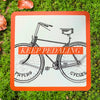 Bright orange card with black illustration of a vintage bicycle and text reading 'Keep Pedaling' overlaid on grass with fallen autumn leaves.