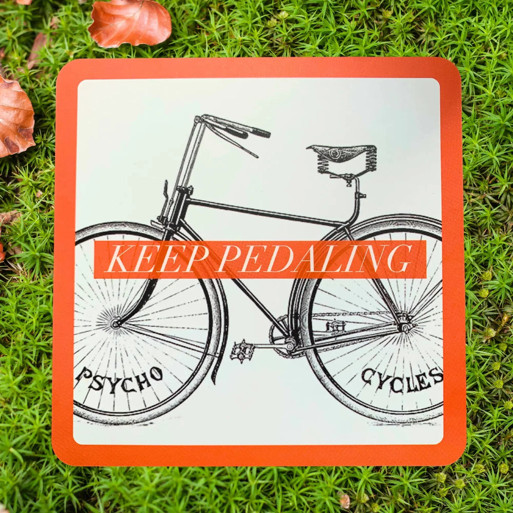 Bright orange card with black illustration of a vintage bicycle and text reading 'Keep Pedaling' overlaid on grass with fallen autumn leaves.
