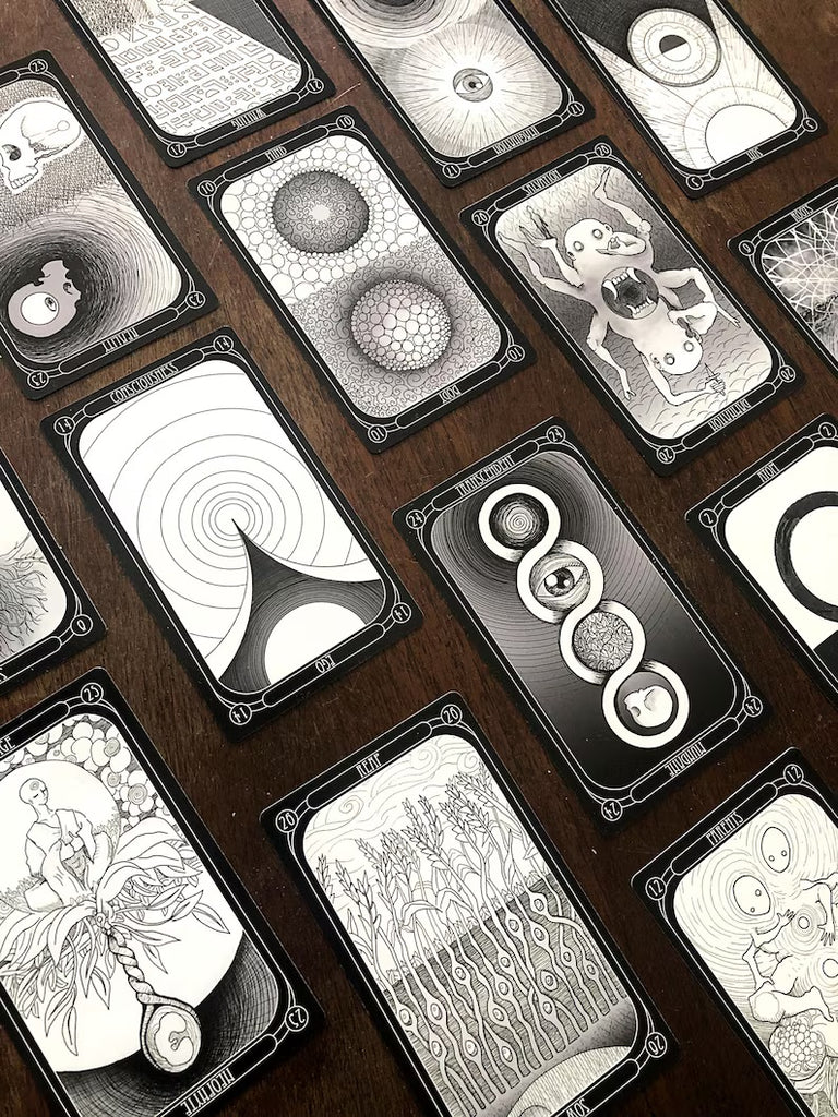 The Eigengrau Oracle Deck Limited Drop by Rage Create is a set of black and white cards featuring surreal, abstract illustrations with organic and geometric designs like plants, eyes, and spirals, arranged on a dark wooden surface.