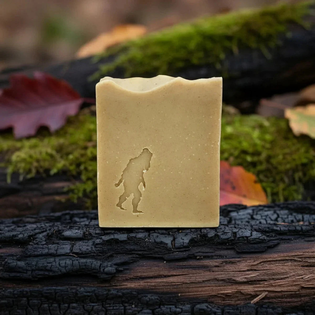 Pale cream-colored soap bar embossed with a Bigfoot silhouette, displayed on a charred log surrounded by vibrant green moss and autumn leaves.