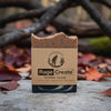 Rage Create coffee scrub soap bar with tan and brown marbled top displayed on concrete among fallen autumn leaves and dark branches, showing the kraft paper label with Bigfoot logo.