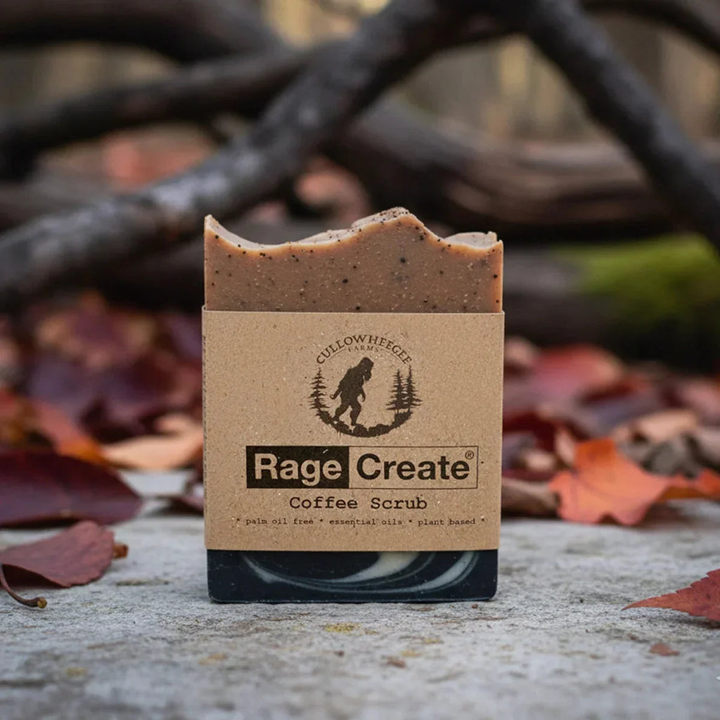 Rage Create coffee scrub soap bar with tan and brown marbled top displayed on concrete among fallen autumn leaves and dark branches, showing the kraft paper label with Bigfoot logo.