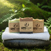 Three Rage Create artisan soap bars in Mountain Air, Coffee Scrub, and Sasquatch varieties displayed on a white towel atop a moss-covered stone, surrounded by lush green ferns in natural daylight.