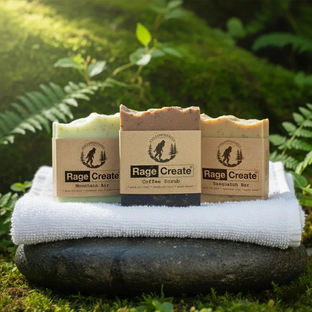 Three Rage Create artisan soap bars in Mountain Air, Coffee Scrub, and Sasquatch varieties displayed on a white towel atop a moss-covered stone, surrounded by lush green ferns in natural daylight.