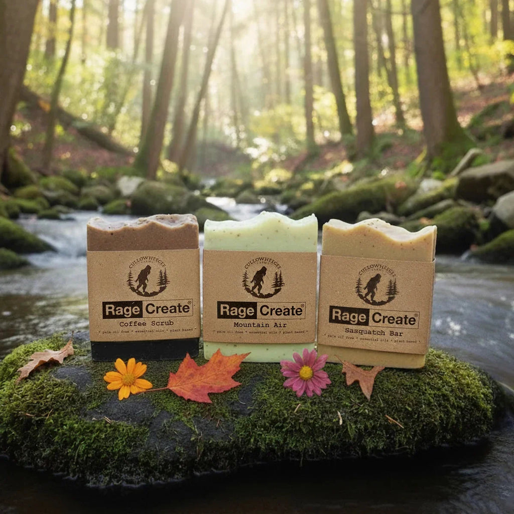 Three Rage Create artisan soaps in kraft packaging displayed on moss-covered stone by a forest stream, with colorful autumn leaves and flowers arranged in front and sunlit trees reflected in the backg