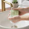 Person washing hands with a green bar soap under flowing water from a brass faucet, with white ceramic sink and potted succulent in background.