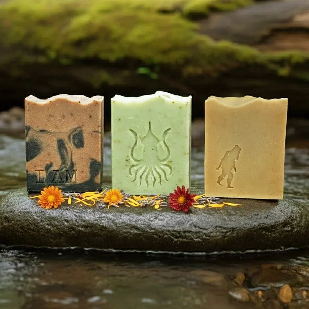 Three artisanal soap bars with embossed designs displayed on a weathered river stone, surrounded by yellow and red flower petals and moss-covered logs in the background.