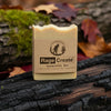 Cream-colored Rage Create Sasquatch Bar soap with kraft paper label sits on charred wood surrounded by bright green moss and autumn leaves in earthy tones.