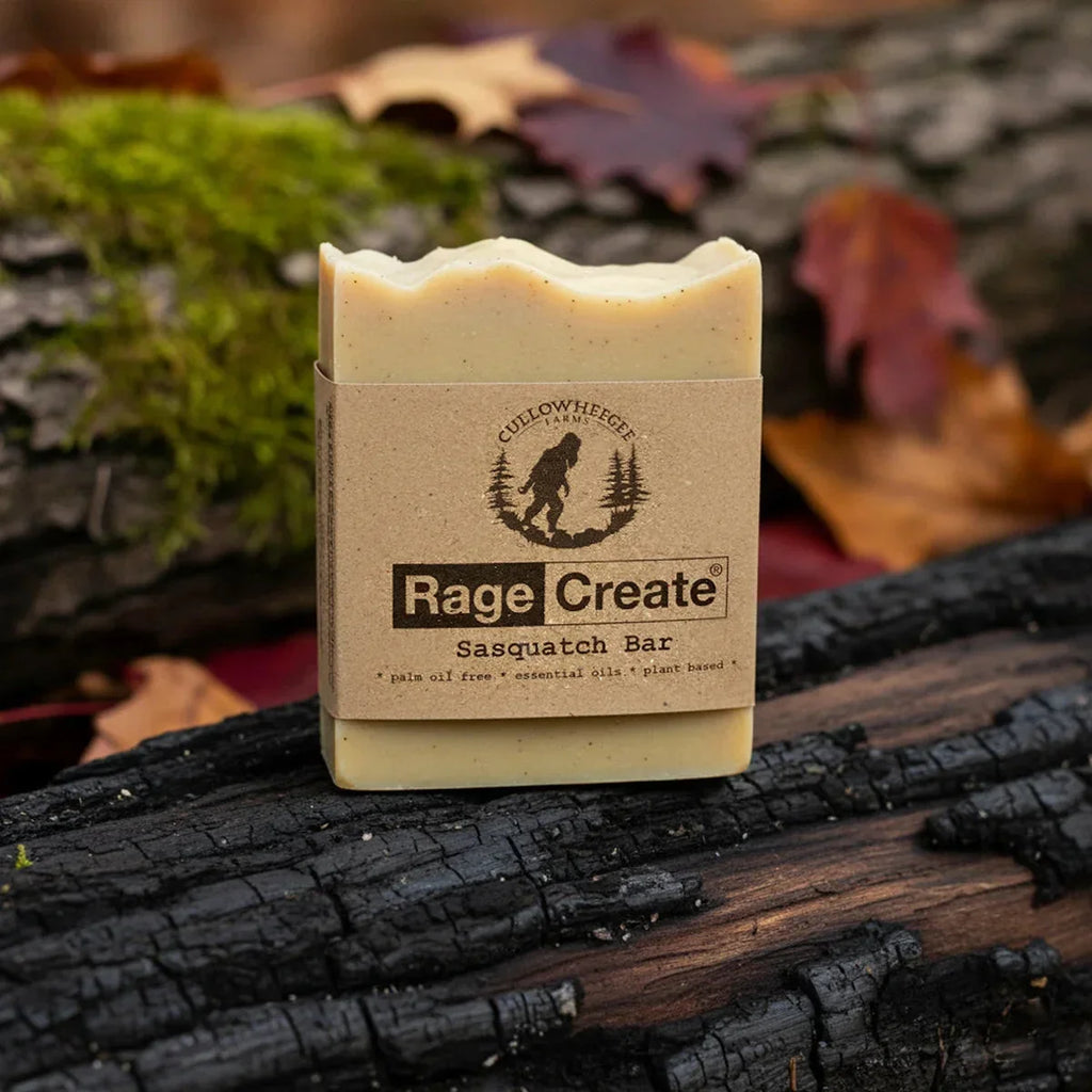 Cream-colored Rage Create Sasquatch Bar soap with kraft paper label sits on charred wood surrounded by bright green moss and autumn leaves in earthy tones.