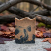 Handmade soap bar with tan and charcoal swirl pattern stamped with branding, displayed on concrete surrounded by autumn leaves and dark branches.