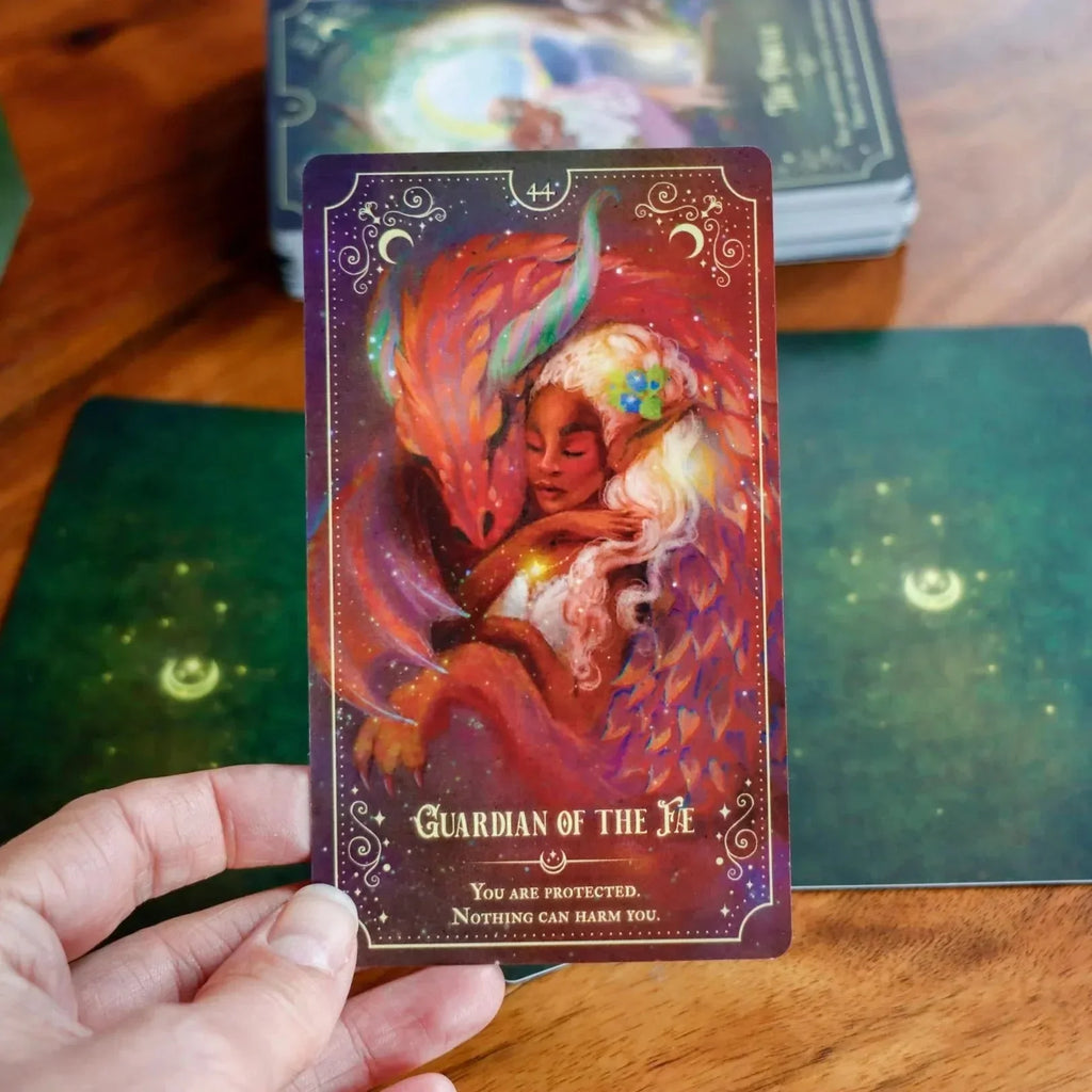 Hand holding the Guardian of the Fire tarot card featuring a fiery-haired woman with flowing red and orange robes surrounded by magical swirling energy and glowing light against a starry purple backgr