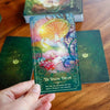 Hand holding The Waking Dream tarot card featuring a glowing golden mushroom and ethereal pink-haired fairy surrounded by vibrant swirling colors and decorative scrollwork against a green deck backdro