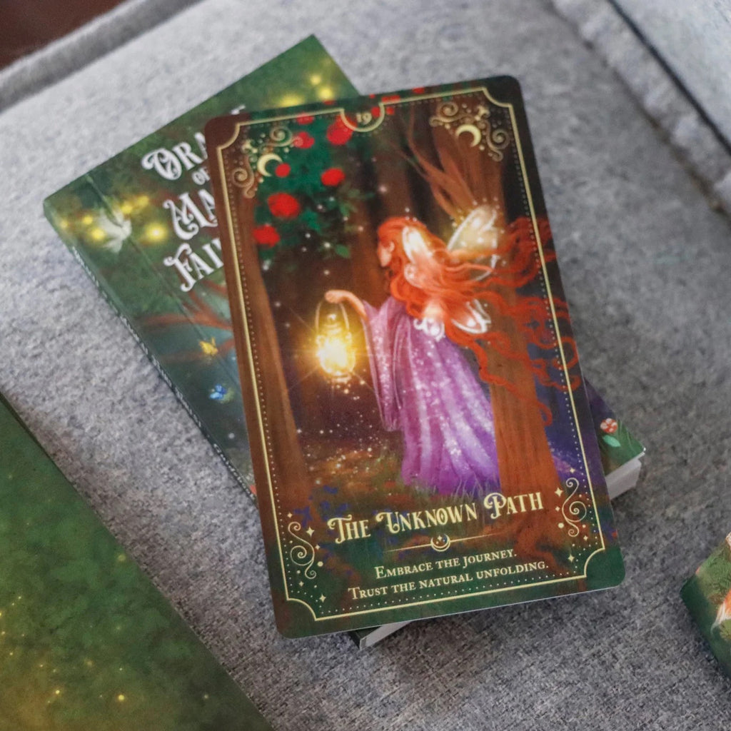 The Oracle of the Magical Fairy Forest Limited Edition by Rage Create features hand-illustrated art of a fairy in purple with a lantern, depicted on The Unknown Path card, displayed atop a closed book on a gray-textured surface.