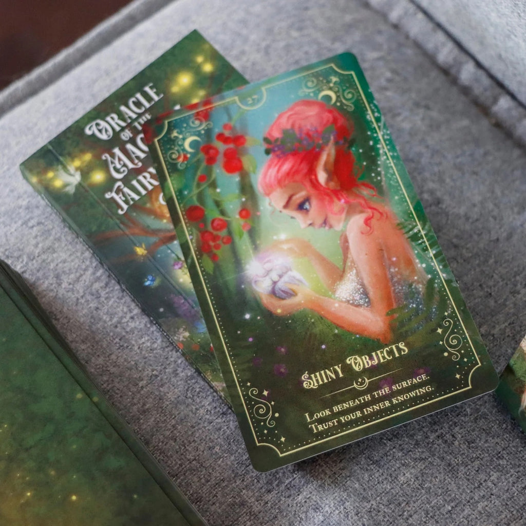 A card from the Oracle of the Magical Fairy Forest Limited Edition by Rage Create shows a red-haired fairy holding a glowing object amid greenery and berries. The hand-illustrated art says: “Shiny Objects. Look beneath the surface. Trust your inner knowing.”.