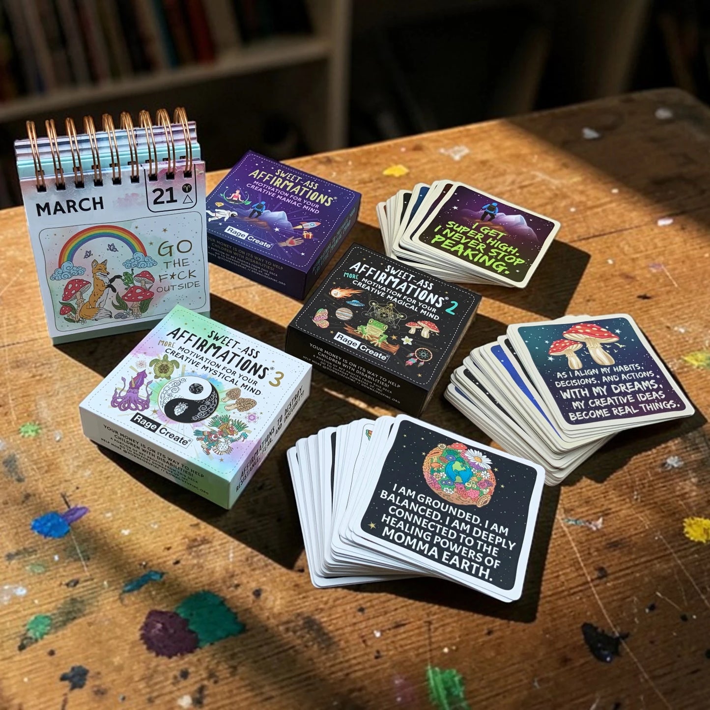 Sweet-Ass Affirmations bundle on a paint-speckled artist's desk — 3 card decks fanned open with readable affirmation cards and the desktop calendar showing March 21