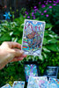 Hand holding the colorful Bison tarot card from The Beasties Oracle Deck against a lush garden backdrop with purple flowers and green foliage.