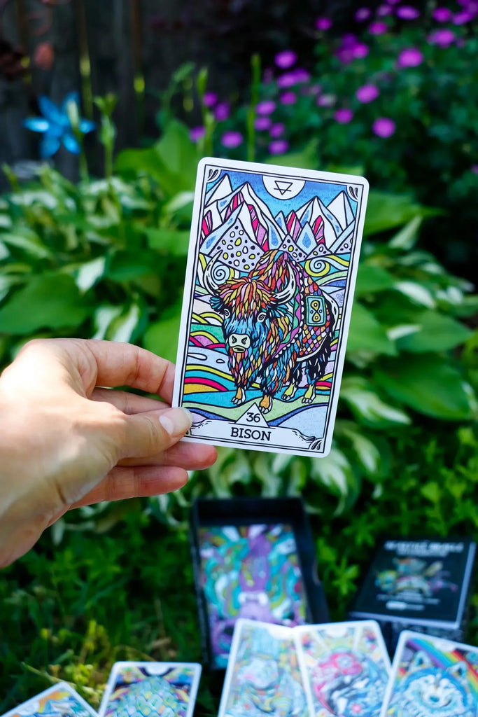 Hand holding the colorful Bison tarot card from The Beasties Oracle Deck against a lush garden backdrop with purple flowers and green foliage.