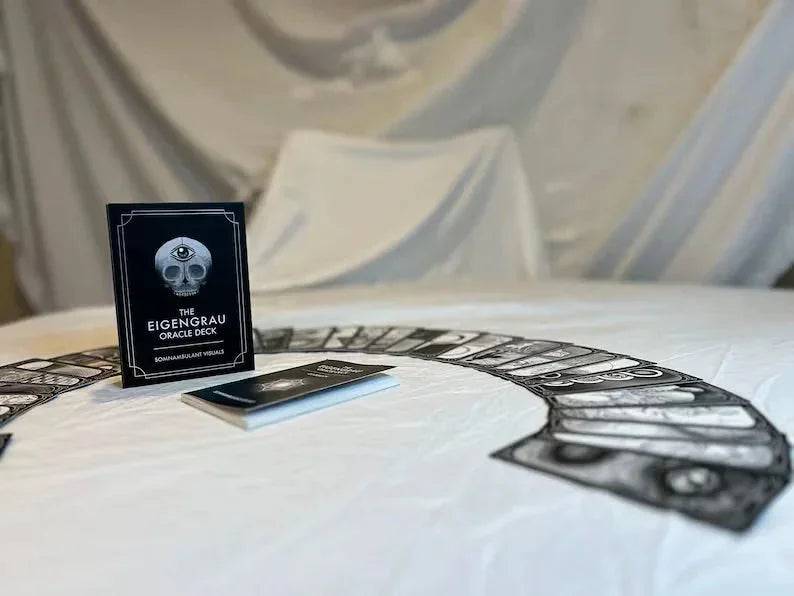 The Eigengrau Oracle Deck black box with silver eye emblem sits upright on white fabric alongside fanned tarot cards, with a cream-colored curtain backdrop.