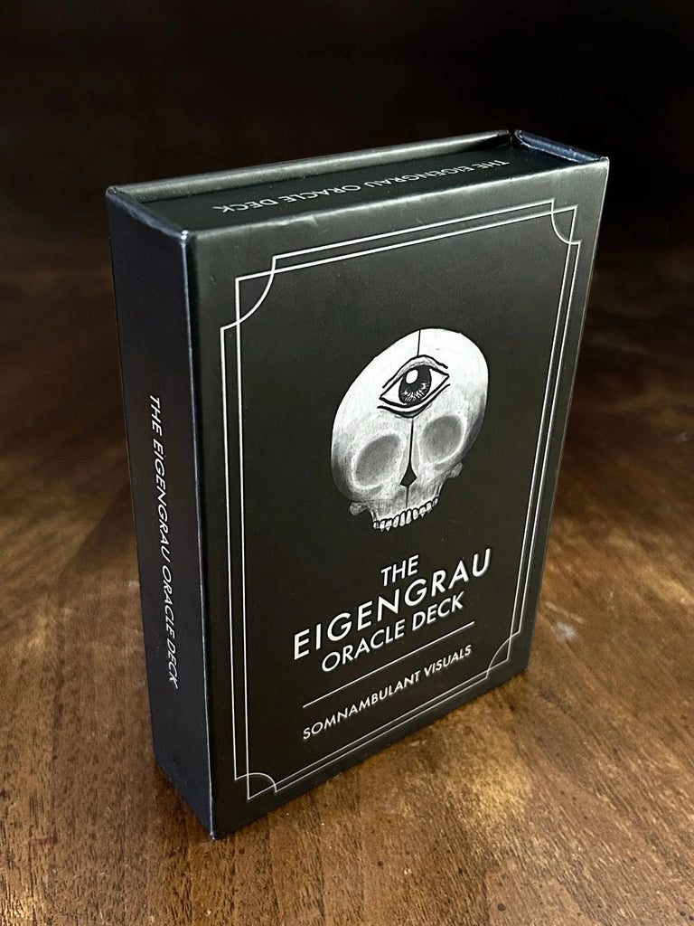 Black tarot deck box with white skull illustration and third eye symbol, displayed at an angle on a weathered wooden table with dark background.