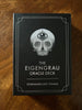 The Eigengrau Oracle Deck box displayed flat on a warm wooden surface, featuring a black cover with a grayscale skull illustration and a third eye symbol, with silver text and decorative corner border